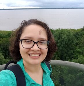 A imagem mostra uma mulher branca, de cabelo na altura dos ombros, de camisa verde e mochila nas costas. Ela está sorrindo. Ao fundo uma paisagem de floresta e de rio.